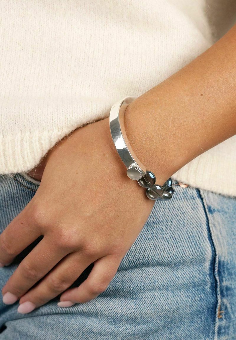 Silverfärgad cuffarmband med en slät yta och rundade svarta pärlor. Bärs på handleden, tillsammans med en krämfärgad stickad tröja och denim.