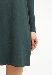 Ciemnozielona sukienka z długim rękawem o miękkiej teksturze, zaokrąglonym dole i lekkim rozszerzeniu w dolnej części. Na boku widoczna ręka.