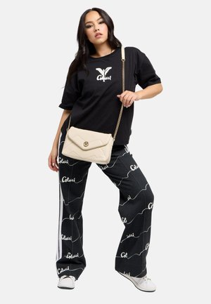 Woman wearing black graphic t-shirt and black pants with white "Colcci" text, white sneakers, and a beige quilted crossbody bag.