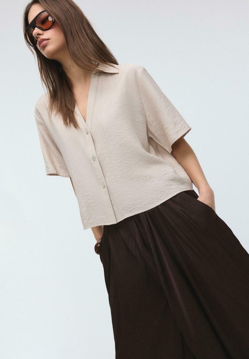 Kvinde iført en oversized beige kortærmet skjorte med knapper, sorte bukser med vide ben og mørke solbriller, står med hænderne i lommerne.