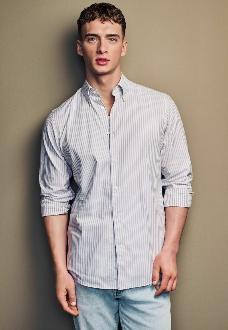 Lichtblauwe en witte gestreepte button-down shirt met lange mouwen, gecombineerd met lichte spijkerbroek, voor een neutrale achtergrond.