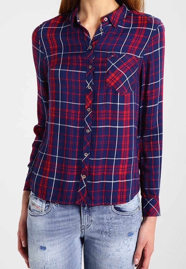 Femme portant une chemise à carreaux rouge et bleu marine boutonnée avec une poche poitrine et un jean bleu clair déchiré.