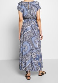 Vestido maxi de estampado azul con mangas tipo capucha, cintura ceñida y detalle de espalda descubierta. La tela presenta diseños de paisley y flores.