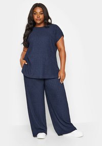 Mörkblå, texturerad topp med korta ärmar, tillsammans med matchande vida byxor; båda har ett subtilt, veckat mönster. Vita slip-on skor.