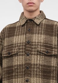 Jeune personne portant une veste en laine à carreaux marron et beige avec des boutons noirs et des poches avant à rabats, visible du cou jusqu'au milieu de la poitrine.