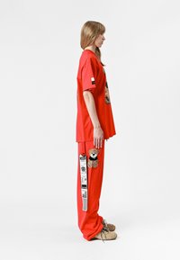 Camiseta roja de gran tamaño con bordado de oso de peluche; pantalones rojos a juego con parche de oso de peluche y detalle de etiqueta. Material de algodón suave, corte relajado.