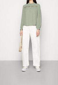 Blusa verde chiaro a maniche lunghe con dettagli plissettati e chiusura con bottoni, abbinata a pantaloni bianchi a gamba larga e sneakers bianche. Tiene una borsa beige.