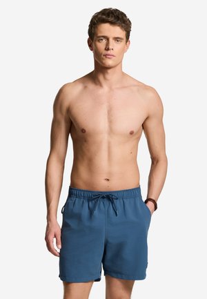 Mladi muškarac s kratkom kovrčavom kosom stoji bez majice, nosi plave kupaće gaće s vezicama, ruke uz tijelo na bijeloj pozadini.