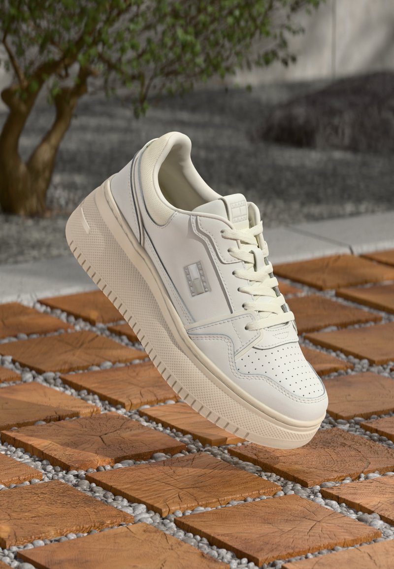 Baskets en cuir blanc à semelle épaisse flottant au-dessus de carreaux de bois carrés à l'extérieur près d'un arbre et de gravier gris.