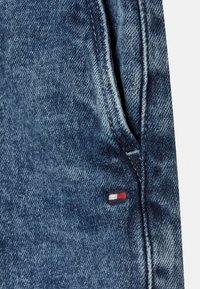 Denim o średnim niebieskim odcieniu, z boczną kieszenią i małym haftowanym logo w kolorze czerwonym, białym i granatowym w pobliżu szwu.