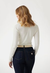 Blusa blanca de manga larga combinada con jeans de mezclilla oscuros. Los jeans tienen un cinturón beige y dos bolsillos traseros con un detalle de logo en forma de triángulo.