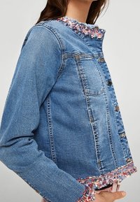 Veste en denim bleu clair au design court. Présente un ourlet effiloché et une bordure texturée et colorée autour du col. Fermeture à boutons.