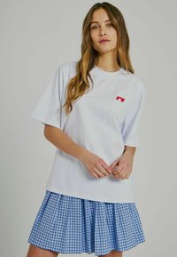 Weisses Baumwoll-T-Shirt mit kurzen Ärmeln und einem kleinen roten Logo auf der Brust, kombiniert mit einem blau-weißen karierten Plissierrock.