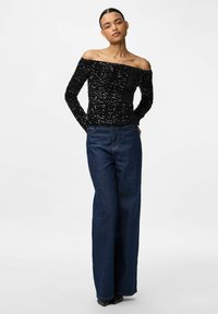 Femme se tenant avec les mains derrière le dos, portant un top à sequins noir sans épaules, un jean large taille haute bleu foncé et des chaussures à talons pointues noires.