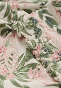 Blomsterdesignat tyg med gröna löv och rosa blommor på en krämfärgad bakgrund, med rynkade och sydda detaljer längs kanterna.
