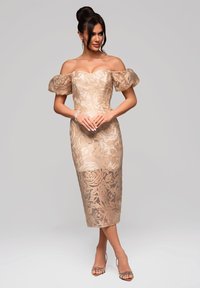Bärande av axlarna beige klänning med blommig spetsdesign, figurnära silhuett, längd till mitten av vadbenet och puffärmar. Kombinerad med remsandaler.
