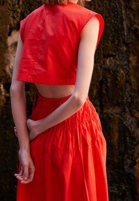 Kvinna i en ärmlös röd crop top och högmidjad röd kjol som står med händerna bakom ryggen framför en stenig bakgrund.