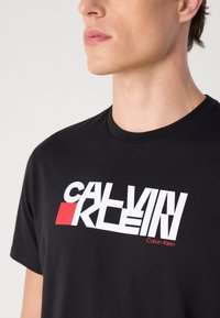 Fekete pamut póló merész fehér és piros Calvin Klein logóval. Kerek nyakú, rövid ujjú, sima textúrával.