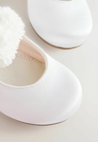 White satin baby shoes with a delicate white fabric flower on the toe, set against a plain light background.