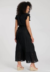 Schwarzes Maxikleid mit Rüschensäumen, strukturiertem Stoff und tiered Saum. Besitzt einen Gürtel in der Taille und Keilsandalen mit Knöchelriemen.