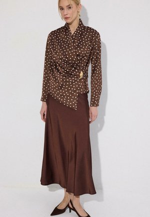 Žena oblečená v hnedej blúzke s bodkami so zlatou brošňou a hnedou saténovou midi sukňou, doplnená nízkymi čiernymi lodičkami.