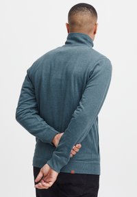 Sweatshirt teal foncé, col montant, manches longues, poignets et ourlet côtelés, avec une petite étiquette logo en bas à gauche.