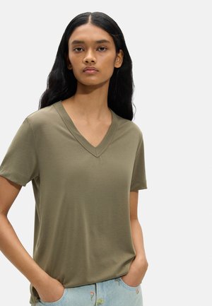 Młoda kobieta z długimi czarnymi włosami, ubrana w oliwkowo zieloną koszulkę z dekoltem w serek i jasnoniebieskie jeansy z kwiatową haftacją, ręce w kieszeniach.