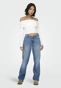 Top blanco sin hombros con detalles fruncidos, combinado con jeans de mezclilla acampanados azul. El conjunto incluye una silueta ajustada y calzado en tonos neutros.