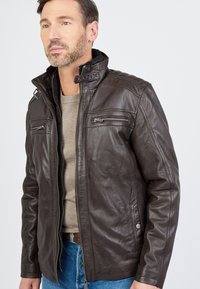 Bruine leren jas met hoge kraag, voorrits en twee ritszakken. Glad oppervlak met subtiele stikselaccenten, gedragen over een beige trui.