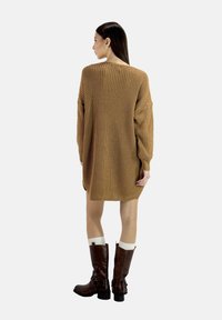 Knit sweater dress in warm brown, featuring oversized sleeves and ribbed texture. Worn with knee-high brown boots and white crew socks.