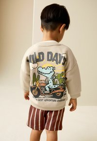 Baltos spalvos džemperis su grafika, vaizduojančia mėlyną krokodilą ant motociklo, apsuptą kaktusų ir tekstu „WILD DAYS“. Ruda dryžuota šortai.