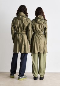 Two individuals wearing olive green, belted, waterproof jackets with hoods. One pairs with dark blue jeans, the other with light green cargo pants.