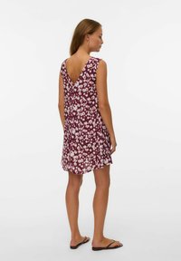Femme debout portant une robe sans manches couleur bordeaux avec un imprimé floral blanc, un dos en V profond, et des tongs noires sur fond blanc.