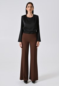 Top lungo a maniche nere in satin abbinato a pantaloni brown a vita alta e gamba larga. La texture liscia e il taglio sartoriale mettono in risalto la silhouette.