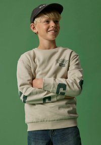 Beige Sweatshirt mit grünen Grafiken und Text auf den Ärmeln, gerippten Bündchen und Saum, kombiniert mit blauen Jeans. Der Hintergrund ist grün.