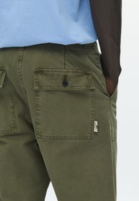 Olivne zelene hlače z žepi spredaj, z gumbno zaporo in tkaninsko etiketo. Material je videti trpežen z mehko teksturo.
