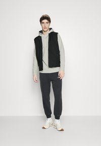 Lys grå hettegenser under en svart vest, kombinert med mørkegrå joggebukser og beige joggesko. Har en avslappet passform og minimalistisk design.