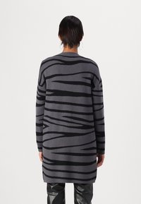 Długi szary kardigan z czarnymi falistymi wzorami, wykonany z miękkiego dzianinowego materiału. Posiada luźny fason i opuszczone ramiona.