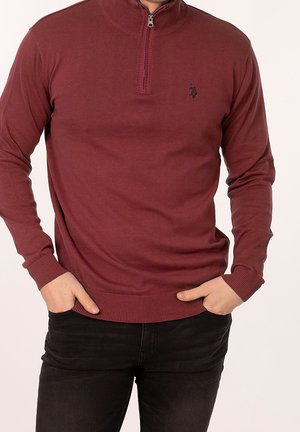 Maroon zip-neck sweater in ribbed texture, featuring a fitted design, long sleeves, and a small logo on the chest, paired with black jeans.