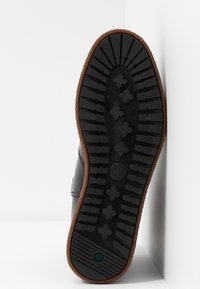 Czarna gumowa podeszwa buta z teksturowanym wzorem bieżnika, z brązowym wykończeniem i logo na środku. Projekt jest funkcjonalny i solidny.