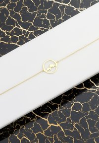 Bracelet enchaîné en or avec une silhouette de montagne circulaire au centre, posé sur une surface d'affichage blanche, contre un fond noir et doré au motif.