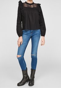 Schwarzes Langarmoberteil mit Rüschenschultern und Spitzeneinsätzen, kombiniert mit blauen Skinny-Jeans mit ausgefransten Knien und schwarzen Stiefeletten.