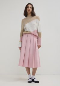 Giovane donna che indossa un maglione beige, bianco e rosa con una gonna midi plissettata rosa, calze bianche e scarpe Mary Jane nere, in piedi contro un muro semplice.