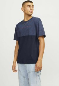 Tamsiai mėlynos spalvos trumpomis rankovėmis marškinėliai su šviesesnio mėlynumo viršutine dalimi ir raudonu užrašu "JACK & JONES". Minkšta medžiaga, laisvas kirpimas ir apvalus apykaklė.
