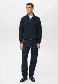 Tmavomodrý sveter s polozipovým golierom, v kombinácii s modrými denimovými džínsami a čiernymi spoločenskými topánkami, nosený stojacim mužským modelom.
