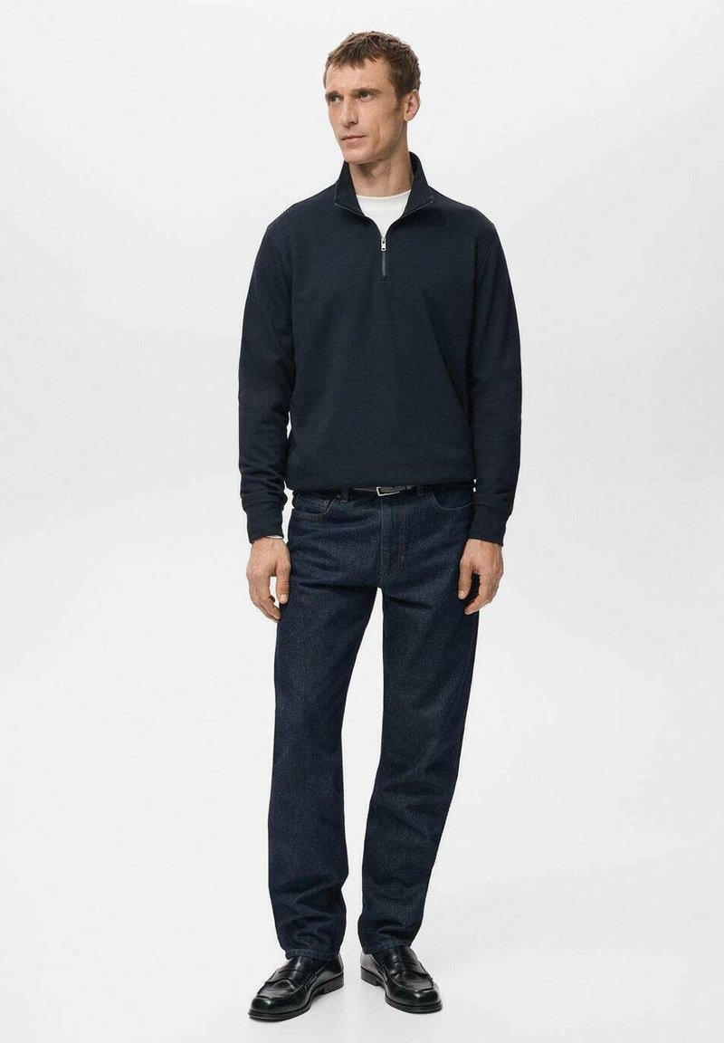 Dunkelblauer Pullover mit Viertelreißverschluss am Kragen, kombiniert mit blauen Jeans und schwarzen eleganten Schuhen, getragen von einem stehenden männlichen Model.