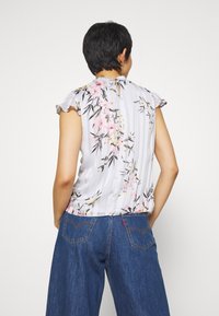 Blus med blommönster i ljusgrått med volangärmar, med en nyckelhålsrygg och ett strukturerat randigt mönster. Kombinerad med vida denimbyxor.