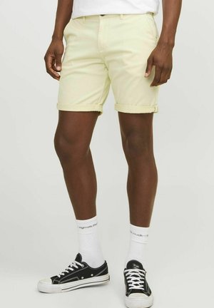 Lysegule bomullsshorts med rulleermer, frontlommer og knappelukking, parret med svarte joggesko og hvite ankelsokker.
