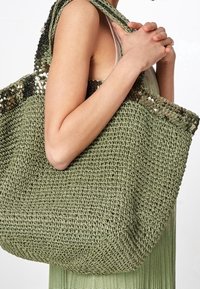 Green crocheted tote bag with a textured finish and a sequined top edge, featuring sturdy straps for carrying.