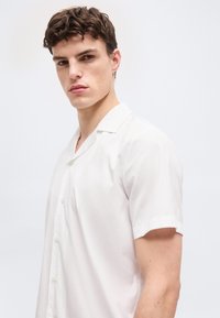 Νεαρός άνδρας με κοντά καστανά μαλλιά, φορώντας λευκό πουκάμισο με κοντά μανίκια και κουμπιά, κοιτάζοντας μπροστά σε φόντο απλό και φωτεινό.